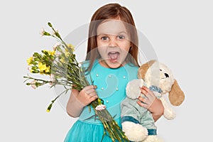 Image of overjoyed blue eyed small child holds her favourite toy and flowers, happy to recieve present on birthday, opens mouth