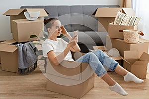 Image of pensive thoughtful woman wearing white t shirt and jeans sitting in a cardboard box and holding cell phone in hands,