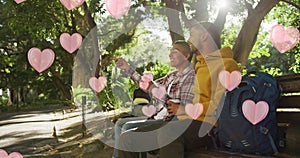 Image of pink hearts floating over happy diverse gay male couple with coffees talking in park