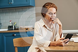 Image of pleased ginger woman using cellphone while siting at table