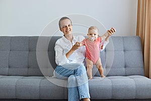 Image of positive optimistic woman wearing white shirt and jeans sitting on sofa with her tiny daughter, baby in pink bodysuit