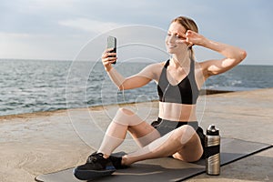 Image of positive woman taking selfie on cellphone and gesturing peace sign while sitting on mat near seaside