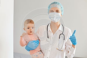 Image of positive young adult female pediatrist wearing medical uniform, holding infant baby girl, examining child`s health in