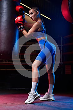 Image of a powerful sportswoman boxer. She is posing near the bag against the background of the ring. The concept of wrestling and