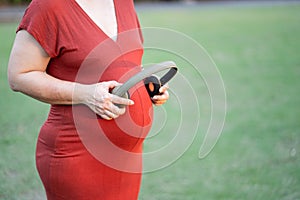 Image of pregnant woman in red maternity clothes standing in garden holding headphone on her baby bump.