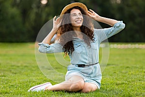 Image of pretty brunette woman wearing straw hat and dress sitting on grass in park