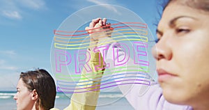 Image of pride flag over diverse lesbian couple holding hands by the sea