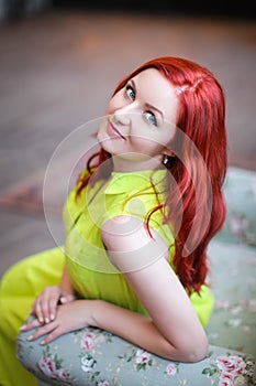 Image of redhead young happy lady in green dress sitting on white armchair on blurred wooden background looking camera