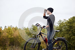 Image of road biker cycling and training on road in forest. Attractive young sportsman stops to have rest after long hours riding