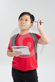 Image of sad smarty boy wearing backpack thinking and touching temple with pen while studying and holding textbooks in hand