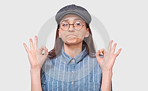Image of satisfied young woman wearing trendy gray cap and round transparent glasses, showing Ok gestures. Pretty student girl