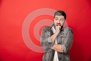 Image of scared man 30s in jeans jacket looking aside on copyspace and biting his fist in horror or fright, isolated over red