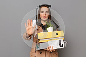 Image of scared shocked woman wearing beige jacket, holding paper folders posing isolated over gray background, showing stop sign