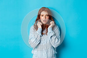 Image of scared teenage girl with red hair, jumping startled and looking alarmed, standing over blue background