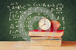 Image of school books on wooden desk, apple and vintage clock over green background with formulas. education concept