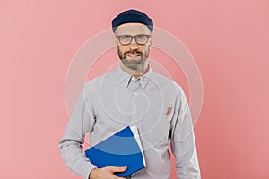 Image of self confident young male with stubble, wears transparent glasses, holds textbook under arm, prepares project work,