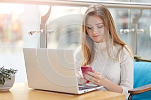 Image of serious busy young girl working online, sitting at cafe, using her laptop and mobile phone simultaneously, having long