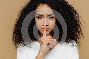 Image of serious confident dark skinned woman makes shush gesture, keeps fore finger over lips, tells secret information, asks not