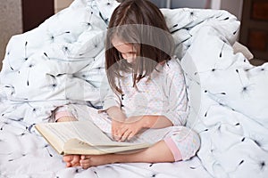 Image of serious preschooler girl reading book in bed, cute kid sitting under blanket with dandelion, darkhaired child reads fairy