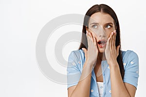 Image of shocked brunette woman gasp, open mouth startled, holding hands on face, staring at upper left corner sale or