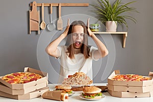 Image of shocked young adult Caucasian woman wearing white casual T-shirt sitting at table in kitchen, screaming with astonishment