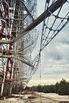 The image shows a large, decaying metal structure in an open area with sparse vegetation, under an overcast sky