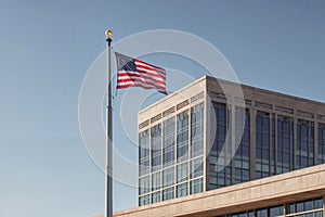 The image shows a modern architectural building with a large American flag waving in the wind in front of it