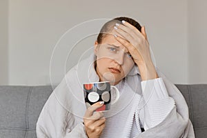 Image of sick ill Caucasian woman being wrapped in blanket sitting on sofa with cup of hot tea, having headache and high