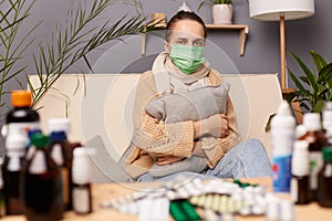 Image of sick unhealthy ill young adult woman wearing warm sweater, scarf and protective mask sitting on sofa in home interior
