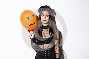 Image of silly beautiful asian woman in witch costume, holding orange balloon with scary face, celebrating halloween