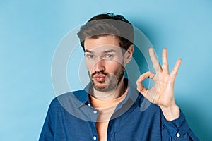 Image of silly young man pucker lips and show okay gesture, praise good thing, compliment excellent choice, standing on