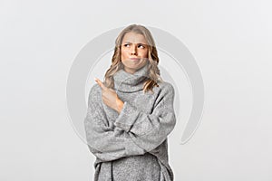 Image of skeptical blond girl in grey sweater, looking indecisive and pointing at upper left corner, smirking doubtful