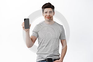 Image of skeptical and disappointed man show empty smartphone screen, frowning and grimacing displeased, standing