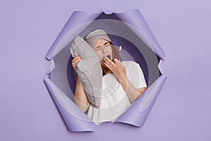 Image of sleepy young girl breaking through paper hole of purple background, holding toothbrush in hand, leaning on pillow, being