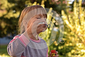 Image of little girl blowing air bubbles with view of green trees and branches behind