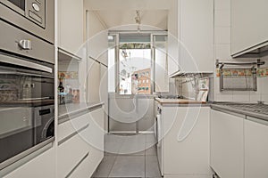 Image of a small kitchen with all the walls covered in white furniture, integrated appliances and a glass and aluminum window in