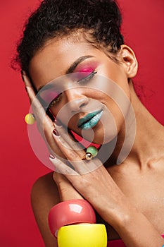 Image of smiling african american woman with bright colorful makeup putting head on palm, isolated over red background