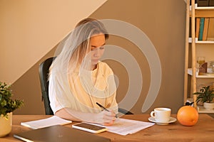 Image of smiling beautiful blond woman writing down notes while sitting at table in office