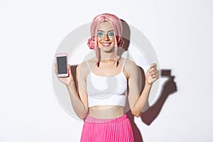 Image of smiling beautiful girl in pink wig, with bright makeup, showing credit card and mobile phone screen, standing