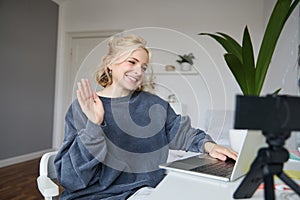 Image of smiling beautiful young woman, chats online on laptop, waves hand, says hello, records video on digital camera