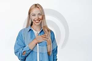 Image of smiling blond girl holds hand on heart and looks flattered at camera, thank you gesture, stands over white