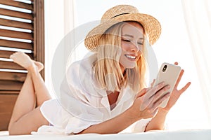 Image of smiling blonde woman using cellphone while lying on bed