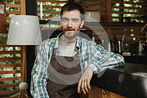 Image of smiling boy wearing apron sitting at the bar while working in cafe or coffeehouse outdoor