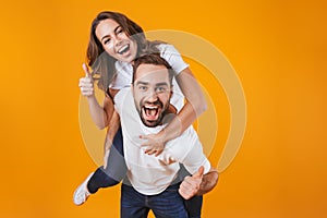 Image of smiling couple having fun while man piggybacking joyful woman, isolated over yellow background