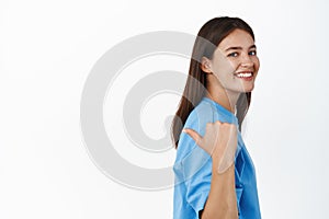 Image of smiling cute healthy girl pointing finger left, showing promo behind her shoulder, on empty space, standing photo
