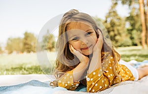 Image of smiling cute little girl lying and relaxing on the blanket at the green grass. Beautiful child taking rest outdoors