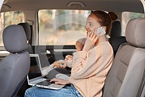 Image of smiling delighted Caucasian woman wearing beige sweater sitting on backseat with her baby and talking smart phone,