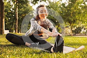 Image of smiling fitness woman 20s wearing headphones working out and stretching legs, while sitting on exercise mat in green park