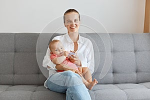 Image of smiling happy woman with bun hairstyle wearing white shirt and jeans, posing with her small daughter, looking at camera