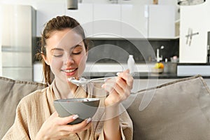 Image of smiling, happy young woman eating breakfast, holding bowl of cereals with milk, having meal at home